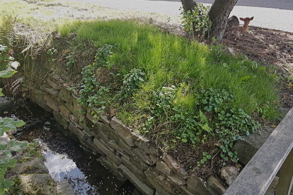 Fertige Natursteinmauer am Bach, die Böschung darüber ist dicht mit Gras und Kräutern bewachsen; rechts ein Holzgeländer, hinten ein Baum mit Rehfigur.