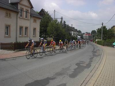 Lang gezogenes Feld von Rennradfahrern in bunten Trikots auf einer Dorfstraße, links ein großes helles Wohnhaus mit Holzzaun, darüber Sommerhimmel.