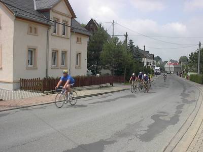 Ein einzelner Rennradfahrer im blauen Trikot fährt voraus, mit Abstand folgt eine kleine Gruppe; links ein helles Wohnhaus, weiter hinten Zuschauer am Straßenrand.